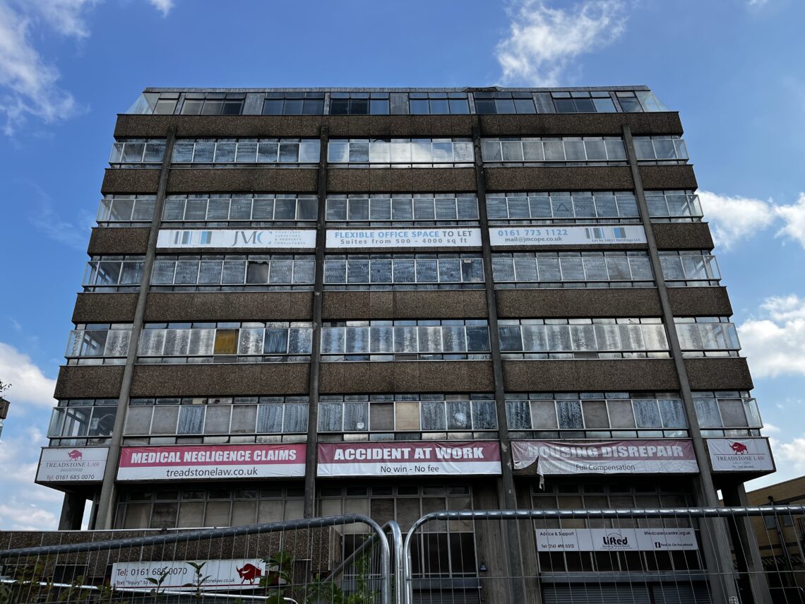 A large, multi-story office building with numerous windows, some dirty or covered. Banners advertise legal services for medical negligence, accident at work, and disrepair. A fence and blue sky with clouds are visible at the bottom and top of the image.