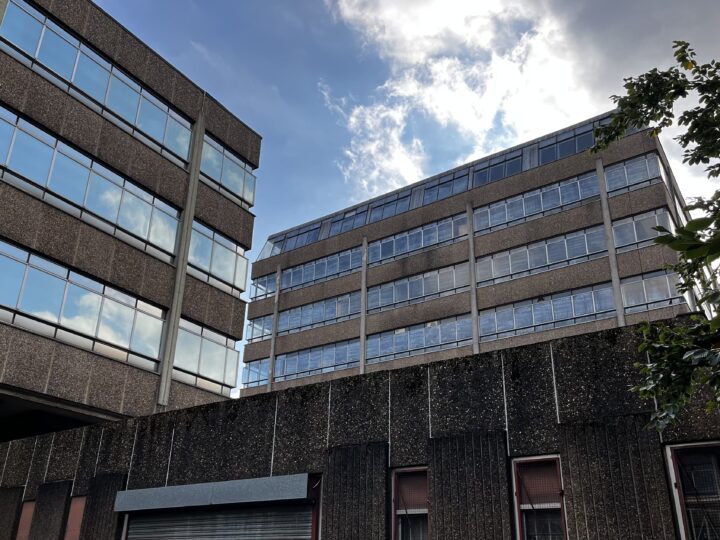 A view of three modern office buildings with rectangular windows and concrete exteriors under a partly cloudy sky. The glass windows reflect the blue sky and clouds. A tree branch enters the frame from the right side.