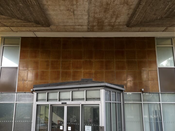 An entrance to a modern building, featuring a glass double door and glass side panels, topped by a flat black canopy. The wall above is covered with square, brown tiles, and the ceiling above is concrete with visible patterns.