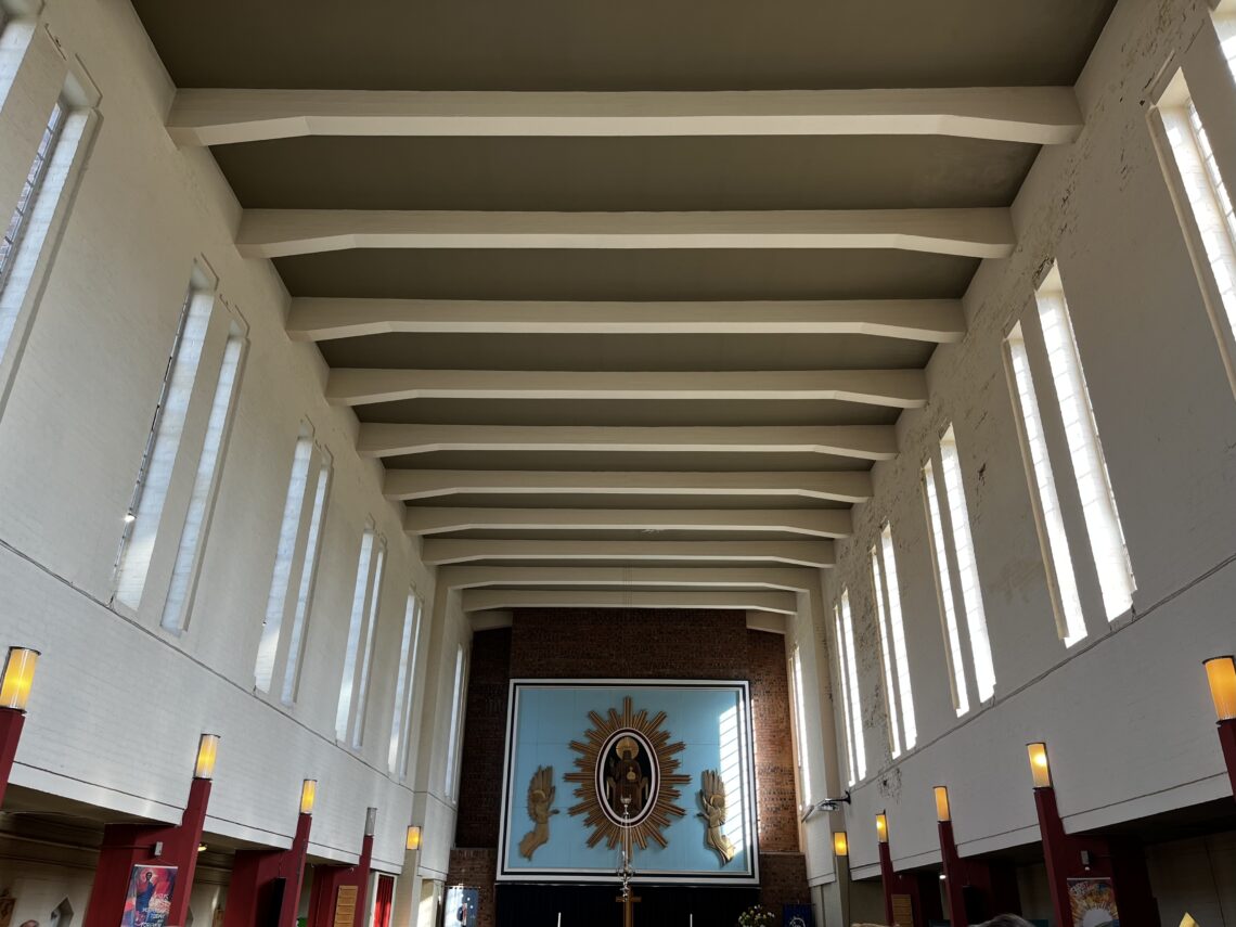 Interior of a modern church with tall white walls, vertical windows letting in natural light, and exposed ceiling beams. At the far end is a large religious artwork featuring a sunburst design and figures, above an altar with candles. Warm lights line the walls.