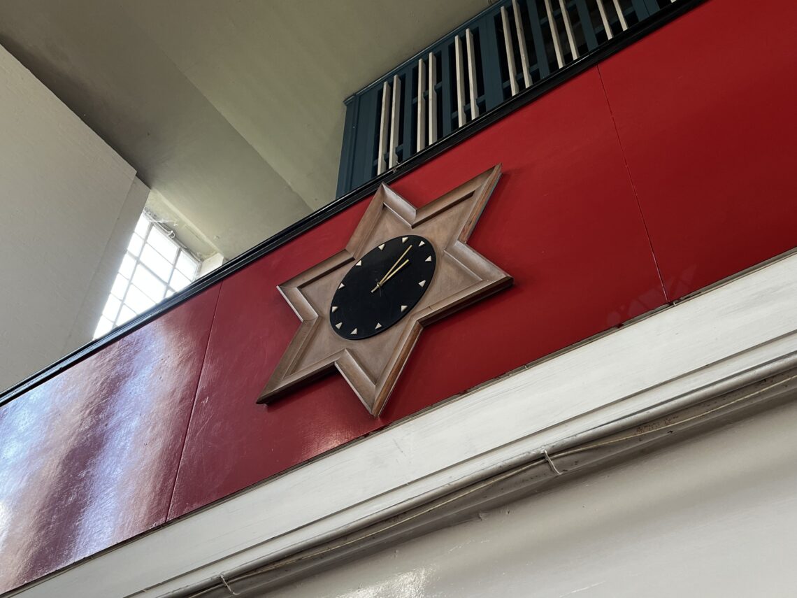 A clock with a black face and gold hands sits in the center of a wooden star-shaped frame mounted on a red wall beneath a railing and near a window, seen from a low angle. The clock shows the time as 12:04.