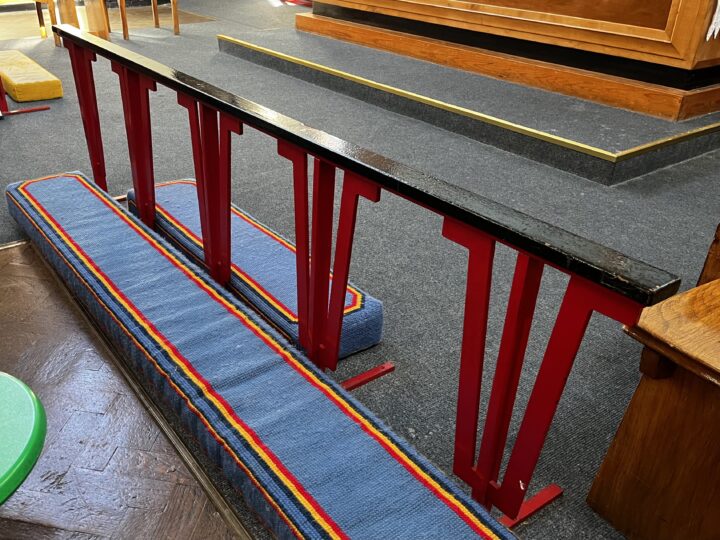 A padded kneeler with blue fabric and rainbow trim sits before a red metal communion rail with a black top inside a church. The rail is on a dark carpeted floor, and wooden steps and platforms are visible in the background.