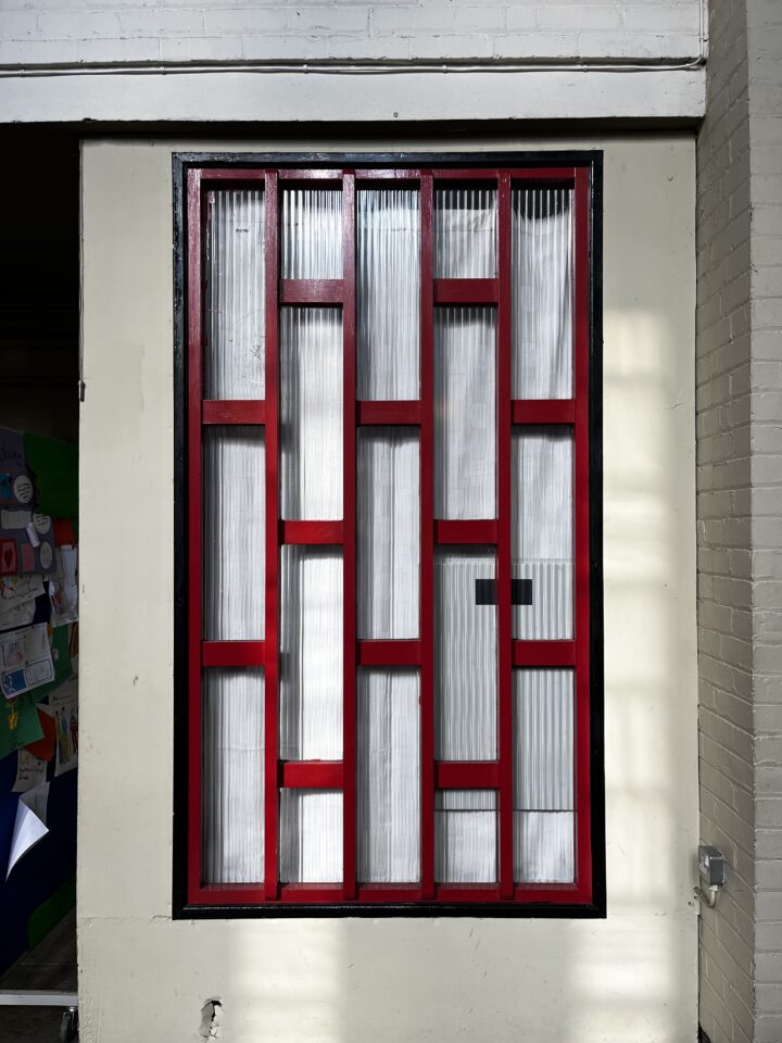 A window with a geometric pattern of red and black frames, featuring vertical and horizontal bars, set in a cream brick wall. The glass panes are translucent with a textured, ribbed surface, letting in diffused light. A dark room is visible to the left.