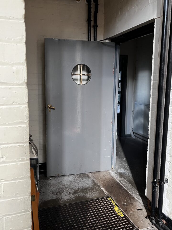 A gray metal door with a round porthole window is partially open in a dim hallway with white brick walls, a black doormat, and visible pipes above. Light shines onto the concrete floor from the doorway, while a radiator is seen in the darker adjoining room.