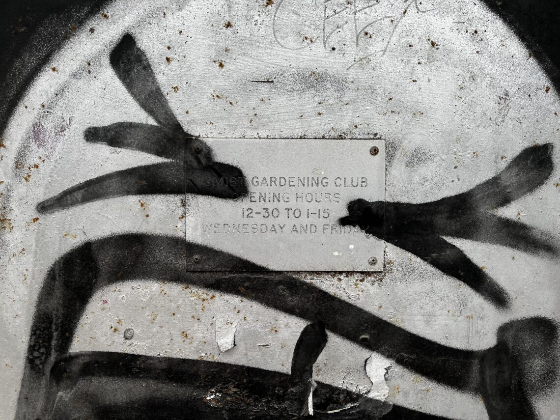 A metal sign with faded, hard-to-read text is mounted on a dirty, weathered surface. Black graffiti in thick strokes obscures much of the sign, but faint text about gardening club opening hours is partially visible behind the spray paint.