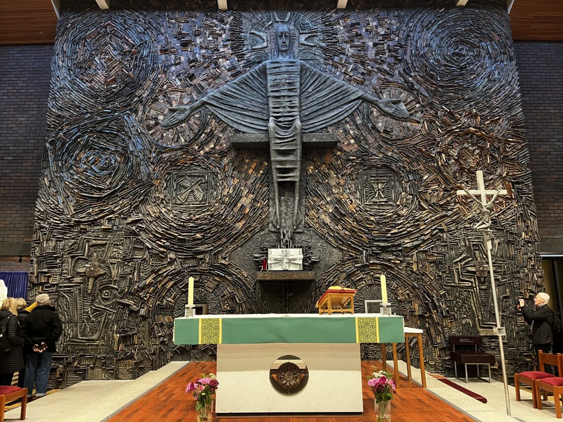 A modern church altar features a large, abstract metal sculpture of Jesus with outstretched arms against a textured, swirling backdrop. The altar is adorned with green cloth, candles, flowers, and a cross. Several people stand or sit near the edges of the scene.