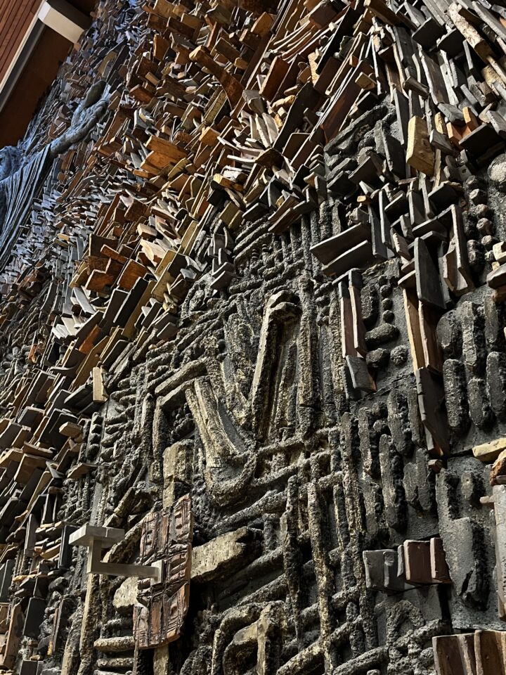 A close-up of an intricate wall sculpture featuring an abstract mix of wood and dark textured materials, with geometric shapes, raised patterns, and protruding wooden blocks arranged in a visually dynamic, layered design. Light casts dramatic shadows, enhancing the depth.