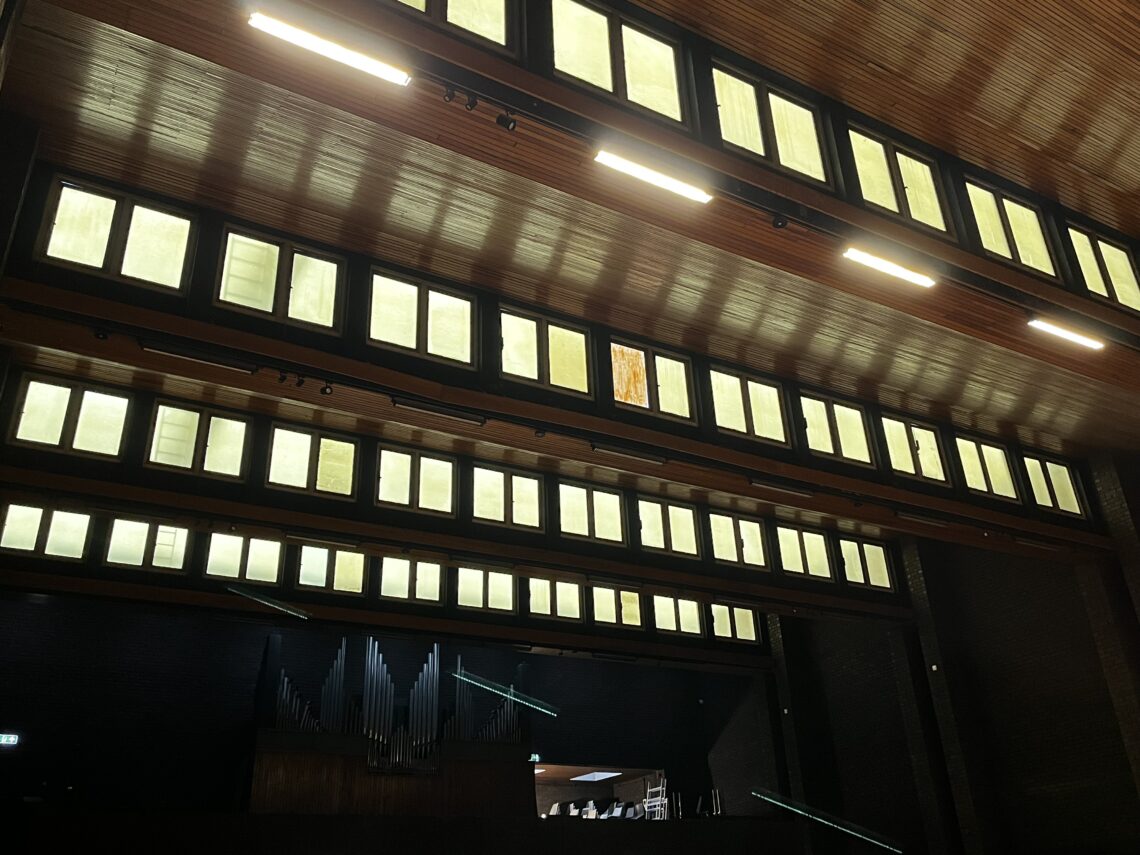 A view inside a dark auditorium showing rows of rectangular, illuminated windows high on the walls. Below, a pipe organ and some chairs are partially visible on a stage, with the ceiling and lighting fixtures overhead.