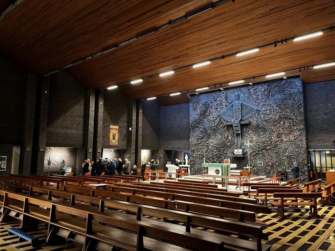 A modern church interior with wooden benches and a high wooden ceiling. People gather near the altar, which stands before a large, textured wall relief featuring abstract designs and a prominent lamb symbol. Warm lighting illuminates the mostly empty, spacious nave.