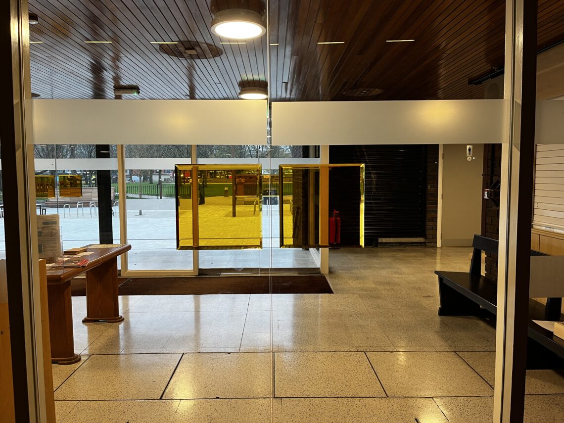 A lobby with terrazzo floor, wooden ceiling, and glass windows. Yellow-tinted glass panels partially cover the windows, with views of an outdoor area and bike racks. A wooden table, a black bench, and a fire extinguisher are visible inside the lobby.