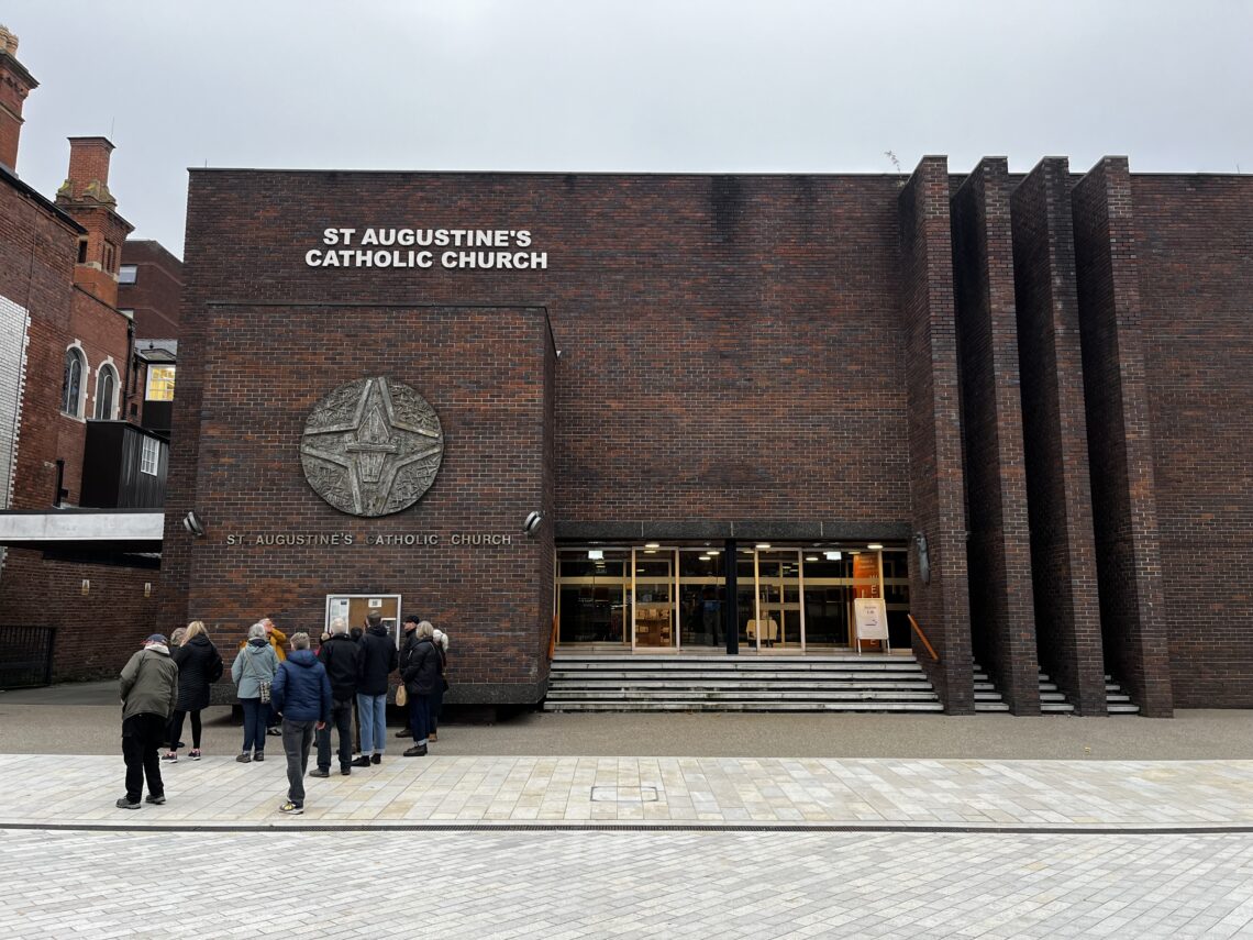 A group of people stand in line outside St Augustine’s Catholic Church, a modern brick building with tall vertical columns, large windows, and a circular religious symbol beside the church name on the exterior wall under a cloudy sky.