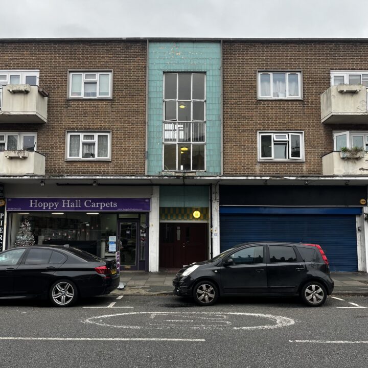 A three-story brick building with two small balconies and turquoise tiles above a central entrance. On the ground floor are a carpet shop with a purple sign and a closed blue shutter. Two cars are parked on the street in front. The sky is overcast.