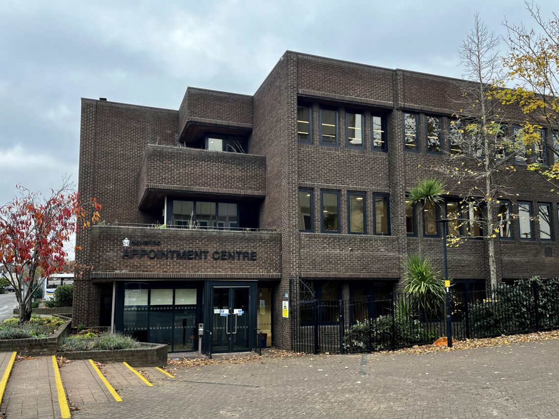 A three-story brown brick building with “APPOINTMENT CENTRE” over the entrance, glass doors, several windows, and balconies. Trees with autumn leaves and green foliage are in front. The sky is cloudy and the area is paved with a ramp and railings.
