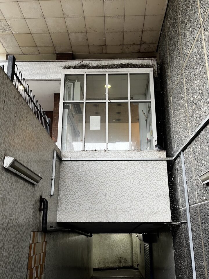 A glass-walled security booth juts out over a dim passageway. The booth sits above ground, supported by concrete, with fluorescent lights and a sign inside. The area below is shadowy, with tile and textured concrete walls and ceilings.