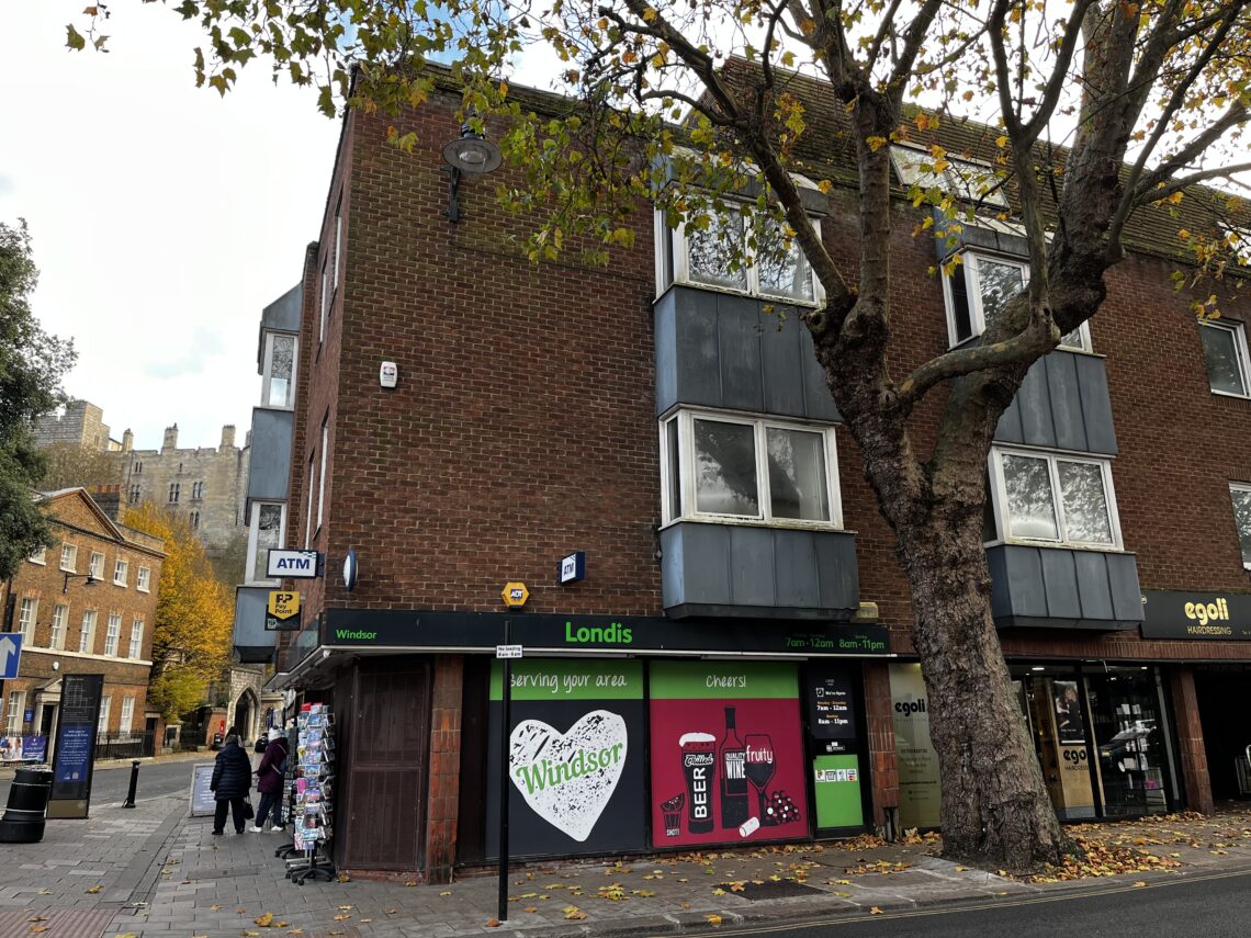 A street corner with a Londis store on the ground floor of a brick building. The shopfront displays colorful ads for drinks and snacks. A large tree stands nearby. People walk past, and an ATM is visible to the left. Autumn leaves are scattered on the ground.