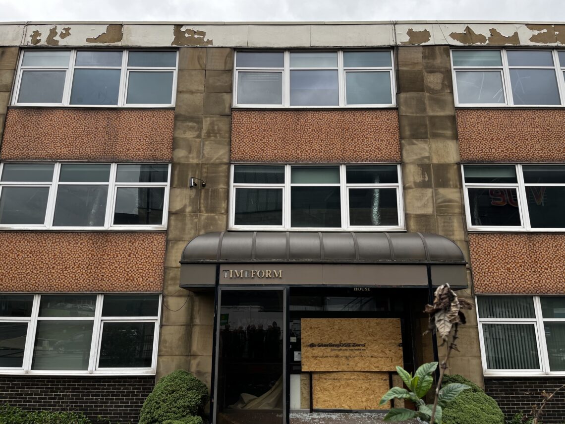 A three-story building with worn, peeling paint on the upper facade, large rectangular windows, and orange mosaic panels. One window is open, and the entrance has a black canopy reading TIMEFORM. The door is boarded up with plywood; some shrubs are in front.