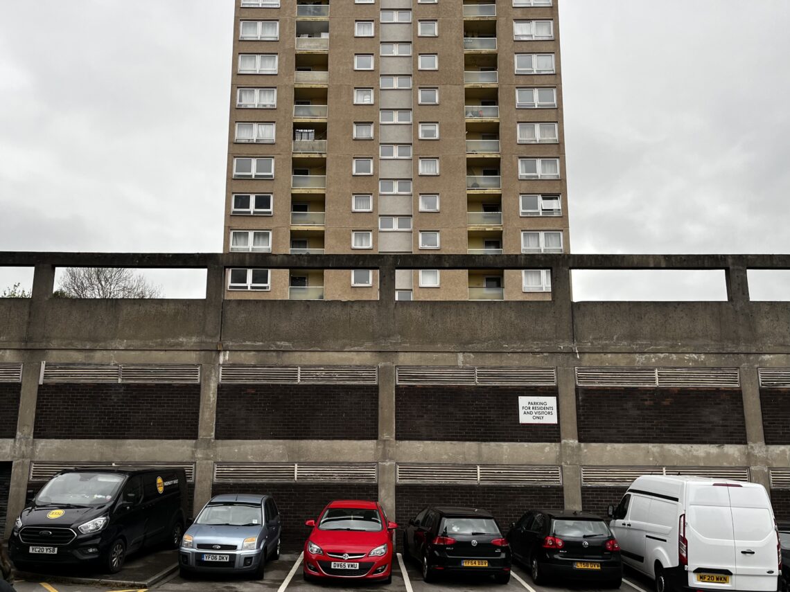 A tall, beige apartment building stands behind a concrete wall. In front, six cars—black, red, and white—are parked in marked spaces. The sky is overcast, and a sign on the wall reads “Parking For Residents Only.”.