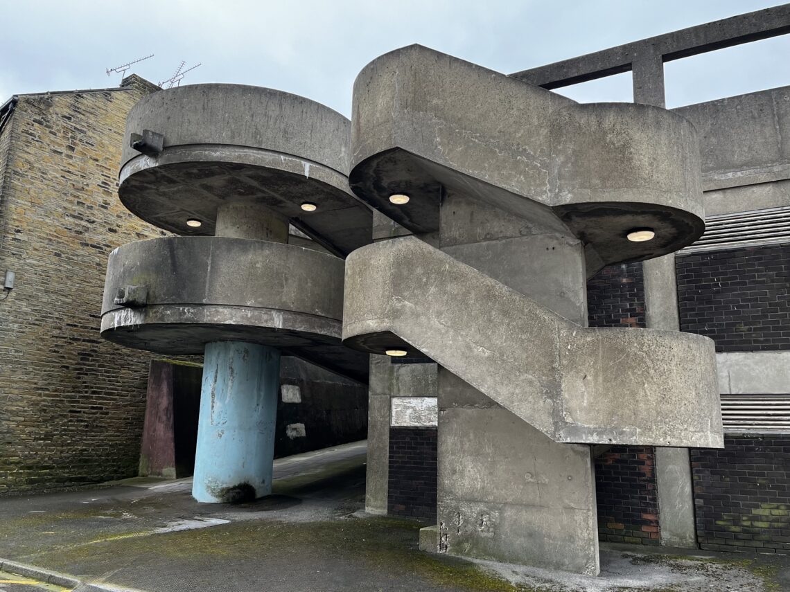 A brutalist concrete spiral staircase with rounded edges and built-in lights juts out from a brick building. A blue column supports part of the stairs. The structure is weathered and sits beside a narrow alley with stone walls. The sky is overcast.