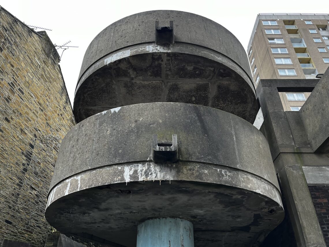 Close-up of a cylindrical, weathered concrete structure with two stacked sections, supported by a blue column. Surrounding it are yellow-brown brick and concrete buildings, with a tall modern apartment block in the background. The sky is overcast.