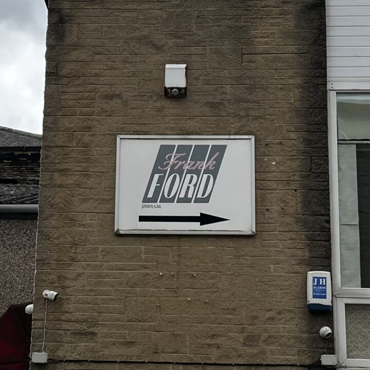 A rectangular sign on a brick wall reads “Frank Ford (2003) Ltd.” with stylized text and diagonal lines. A black right-pointing arrow is below the text. There is a security light above the sign and a blue alarm box mounted to the right.