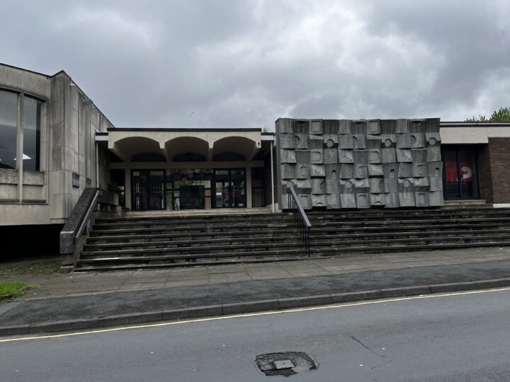 A modern concrete building features wide steps leading to glass entrance doors. On the right, a large abstract gray relief decorates the facade. The sky is overcast, and a wet street runs in front of the building.