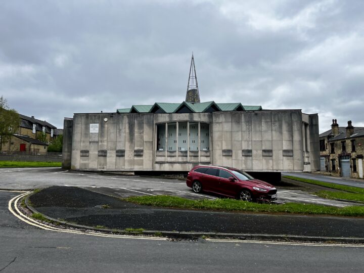 A modernist concrete church with zigzag roof edges and a tall spire stands on a cloudy day. A red station wagon is parked in front. The building is surrounded by empty pavement and bordered by old stone houses. The scene appears quiet and overcast.
