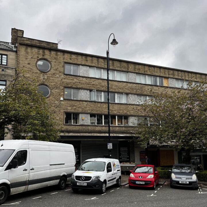 A three-story stone building with round and rectangular windows stands behind parked vehicles, including a white van, a red car, a small silver car, and another white van. Overcast sky and leafy trees partially obscure the building’s lower level.