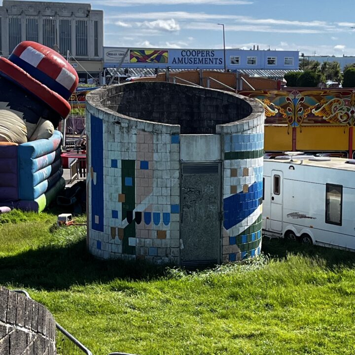 A circular, tiled concrete structure with a metal door stands on grass. Its blue and green mosaic tiles are faded. Behind it are a white camper, a yellow amusement ride, a deflated inflatable hat, and a building labeled COOPERS AMUSEMENTS.