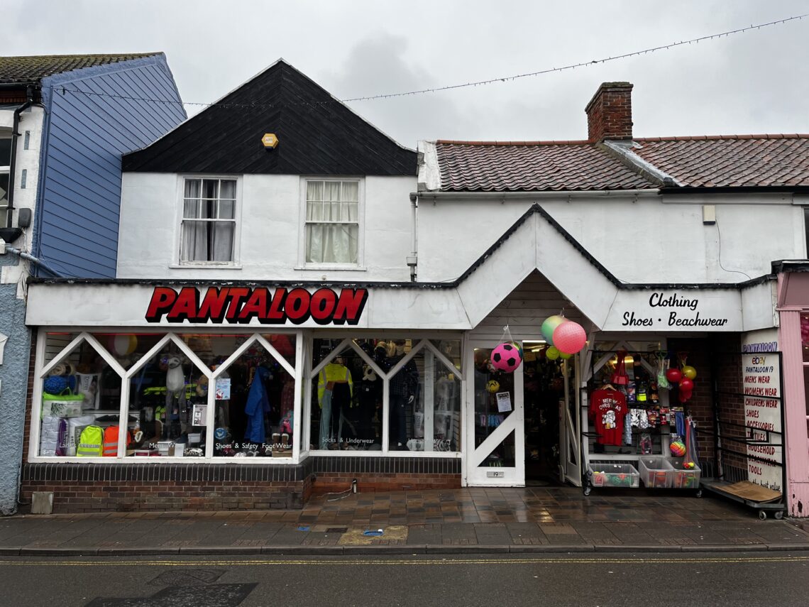 A street view of two neighboring shops. The left store, “PANTALOON,” displays clothes in its large window. The right shop, labeled “Clothing Shoes Beachwear,” shows colorful items and hanging decorations at the entrance. The pavement is wet, and the sky is cloudy.