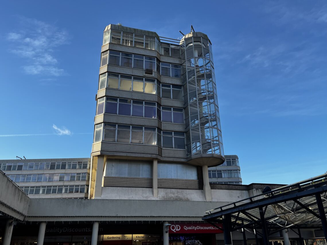 A modernist building with angular lines and a glass cylindrical stairwell rises above street level, catching sunlight. Below, shopfronts including Quality Discounts are visible. The sky is clear and blue, with a few wispy clouds overhead.