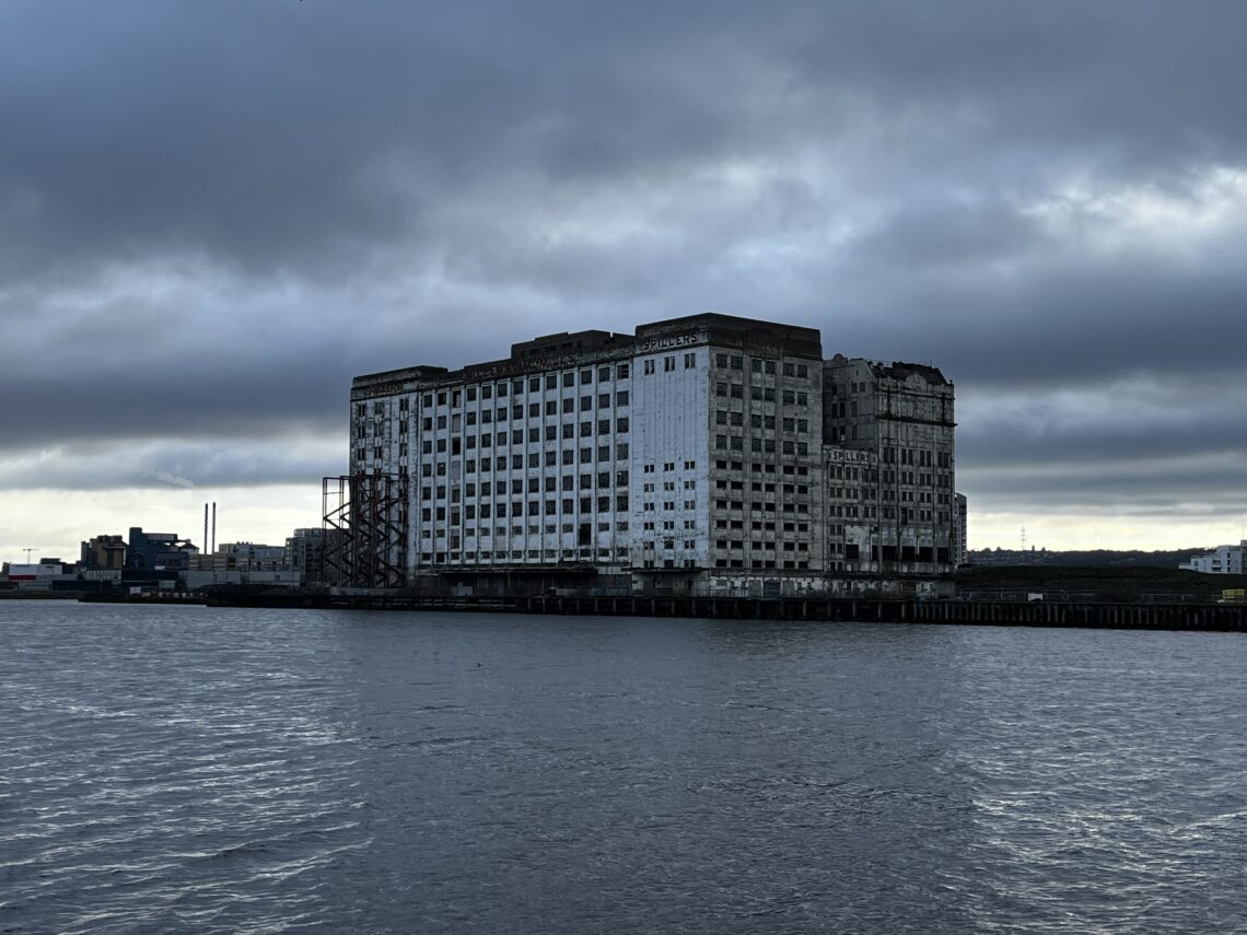 A large, abandoned white building with broken windows stands on a waterfront under a cloudy, overcast sky. The water is calm, and industrial structures are visible in the distant background. The building appears weathered and partly deteriorated.