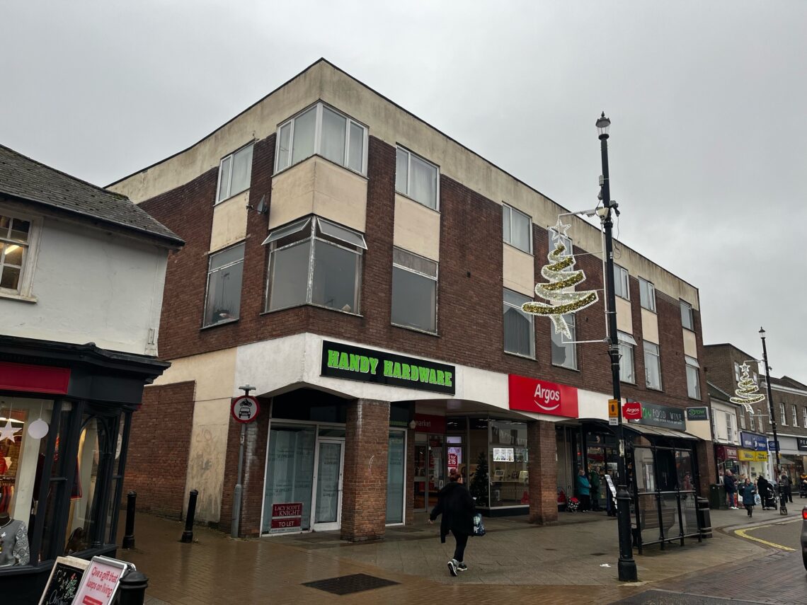 A street scene features a three-story corner building with brown brick and white panels. Shops include Handy Hardware and Argos. People walk by on a wet, cloudy day. Festive tree lights hang from lamp posts, and nearby stores are visible on either side.
