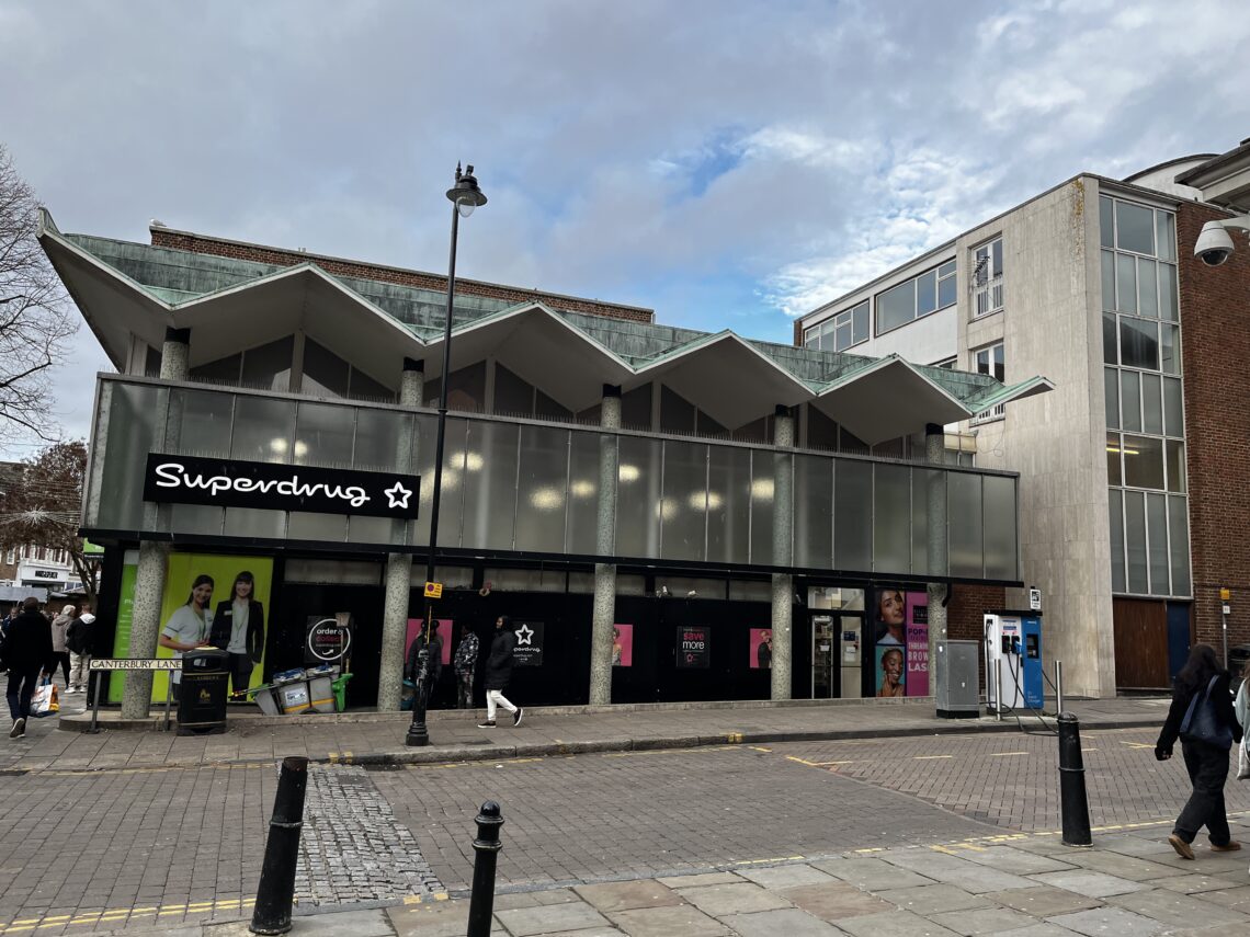 A modern Superdrug store with large windows and a zigzag roof sits on a quiet city street. People walk past, and a bicycle is parked nearby. The sky is partly cloudy, and adjacent buildings have concrete and brick exteriors.