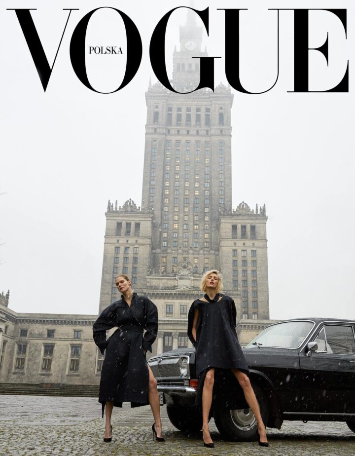 Two women in stylish black coats and high heels pose confidently by a vintage black car, with one leg forward. They stand in front of the towering Palace of Culture and Science in Warsaw on a gray, snowy day. The words “VOGUE Polska” are at the top.