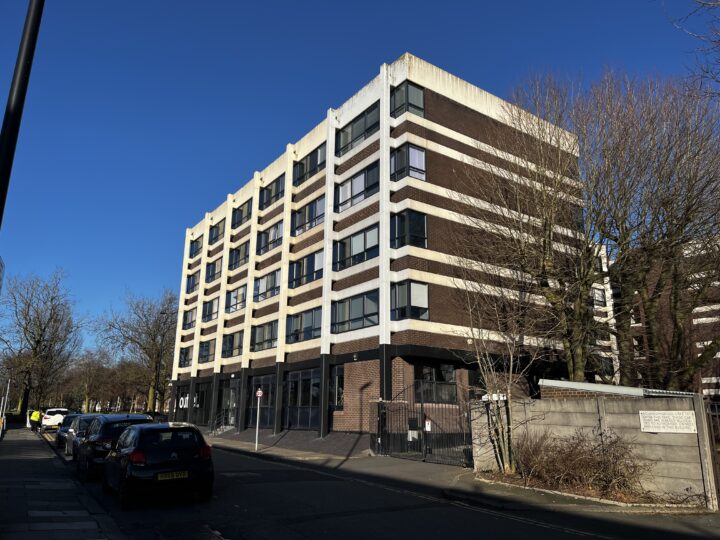 A five-story rectangular building with brown and white horizontal stripes stands on a sunny street. Bare trees line the sidewalk, and several parked cars are visible. The sky is clear blue, and a fenced area is in the foreground on the right.