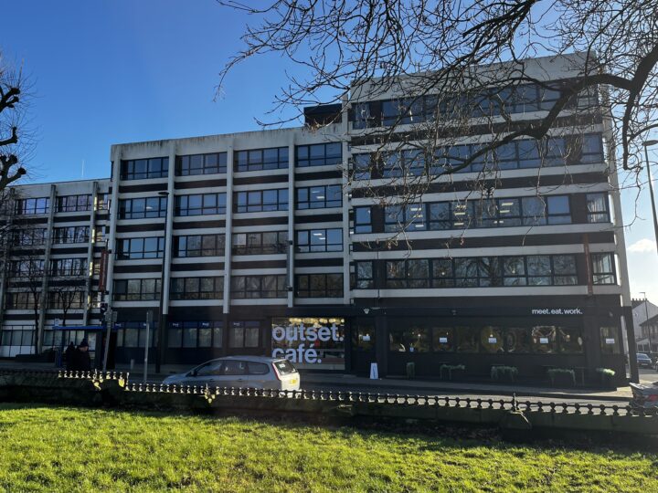 A large, modern four-story office building with many windows. The ground floor has a Outset Café with tables outside and a sign reading meet. eat. work. A few cars are parked in front, and leafless trees and green grass are visible in the foreground.