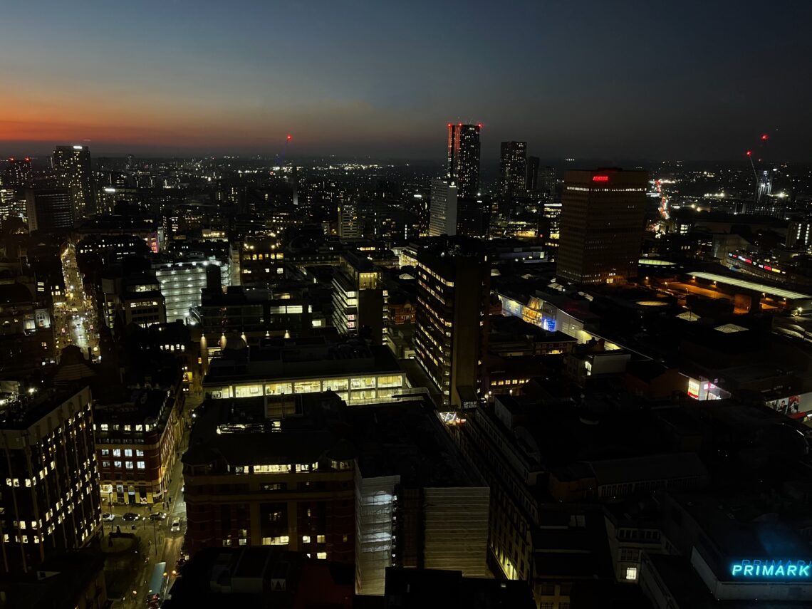 A cityscape at dusk with illuminated buildings and streets. The sky transitions from orange at the horizon to deep blue above. Several high-rise buildings have glowing windows; a blue “PRIMARK” sign is visible in the lower right. Red lights mark taller towers.