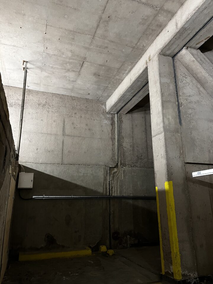 A dimly lit concrete parking garage corner with exposed pipes and a small utility box on the wall. Yellow paint marks the edges of columns and curbs, and shadows fall across the rough, gray concrete surfaces.