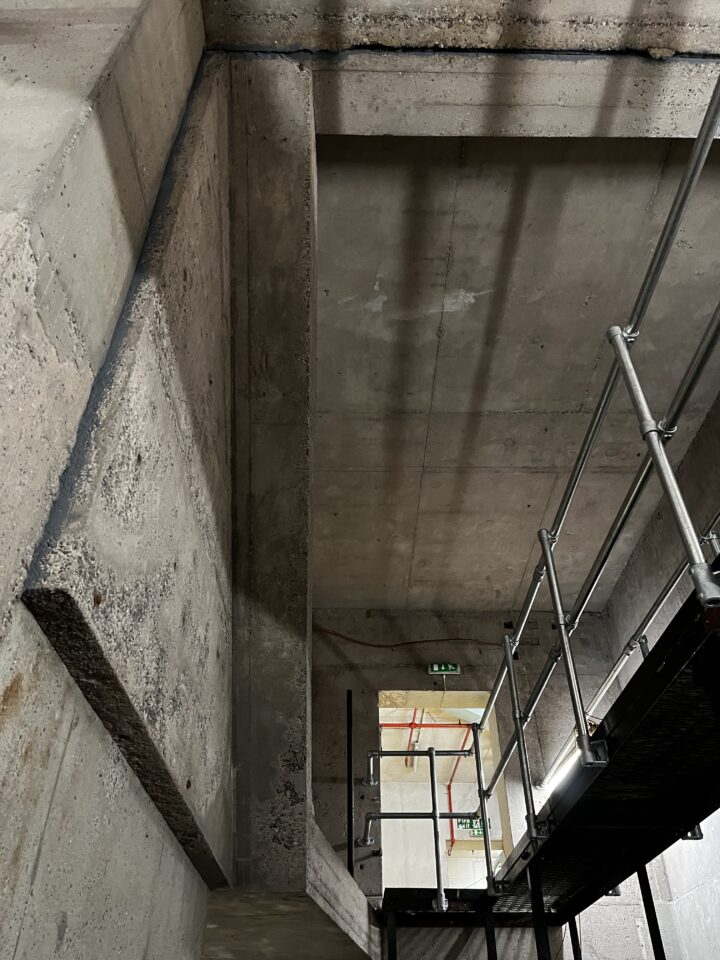 A view looking up a concrete stairwell with visible stairs, walls, and ceiling. Metal scaffolding stands on the right, partly blocking the lower stairs. Lighting creates shadows, and there are green exit signs and orange pipes visible in the background.