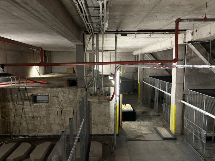 Dimly lit industrial basement with exposed concrete walls and ceilings. Red pipes run overhead; a bar with hazard tape hangs across the top right. Metal fencing and yellow posts form corridors below, and construction materials are scattered around.