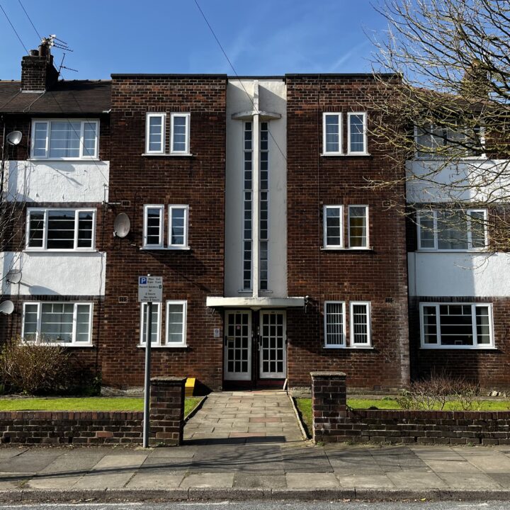 A three-story brick apartment building with white accents and a central entrance. There are multiple windows on each floor, some with satellite dishes. A small front lawn, paved path, low brick wall, and a street sign are visible in the foreground under a clear sky.
