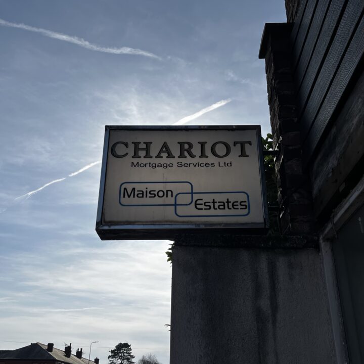A rectangular sign reads “CHARIOT Mortgage Services Ltd” above the “Maison Estates” logo, attached to a corner building. The background sky is blue with light clouds and contrails, and rooftops and a tree are visible in the lower left corner.