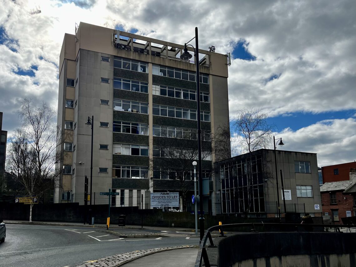 A tall, beige and gray office building with many windows stands on a city corner under a cloudy sky with patches of blue. A To Let sign is visible on the buildings lower facade, and leafless trees and streetlights surround the scene.