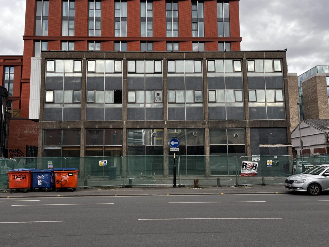 A grey, four-story, vacant building with boarded windows, some broken glass, and protective green fencing around the base. Two red dumpsters sit at the left. A silver car is parked on the right. Overcast sky and a tall red-brick building behind.