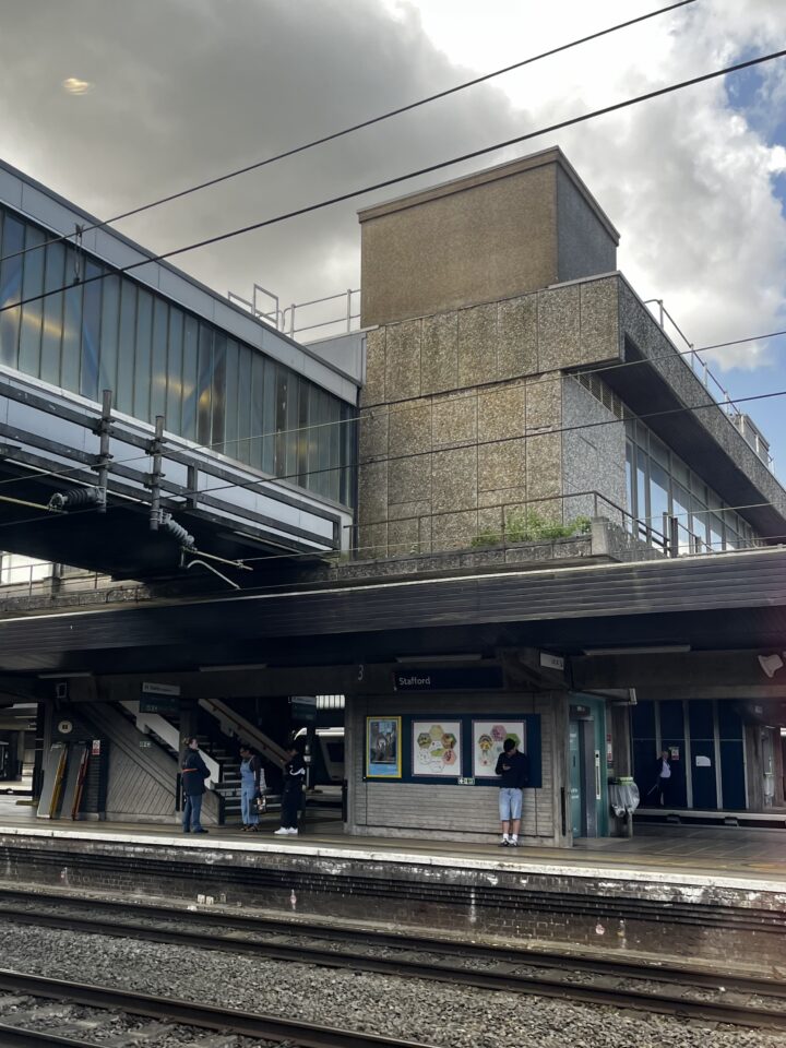 Image of 1950's brutalist train station, with the concrete panel covered lift shaft visible in the centre, and an adjoining glass tunnel above rail level. Also visible is a staircase with diagonal concrete panelling.
