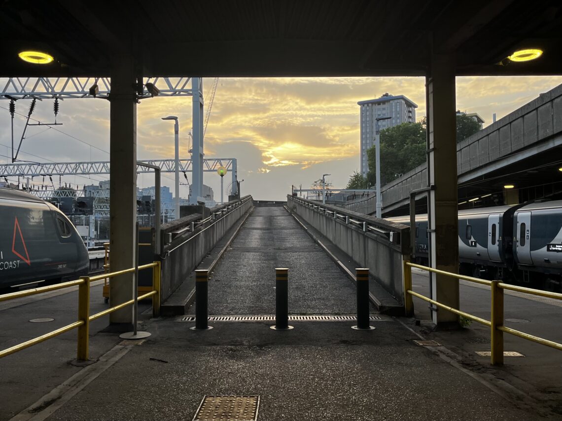 A ramp with short sides made of concrete. In the sky, the sun is breaking through the clouds. There are trains either way of the ramp, as well as other central London buildings in the background.
