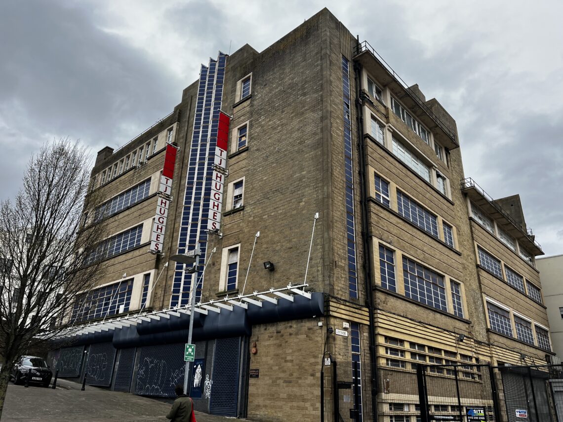 A large, semi-derelict art deco building made from brown brick. Featuring TJ Hughes branding, and all windows blacked out in navy.