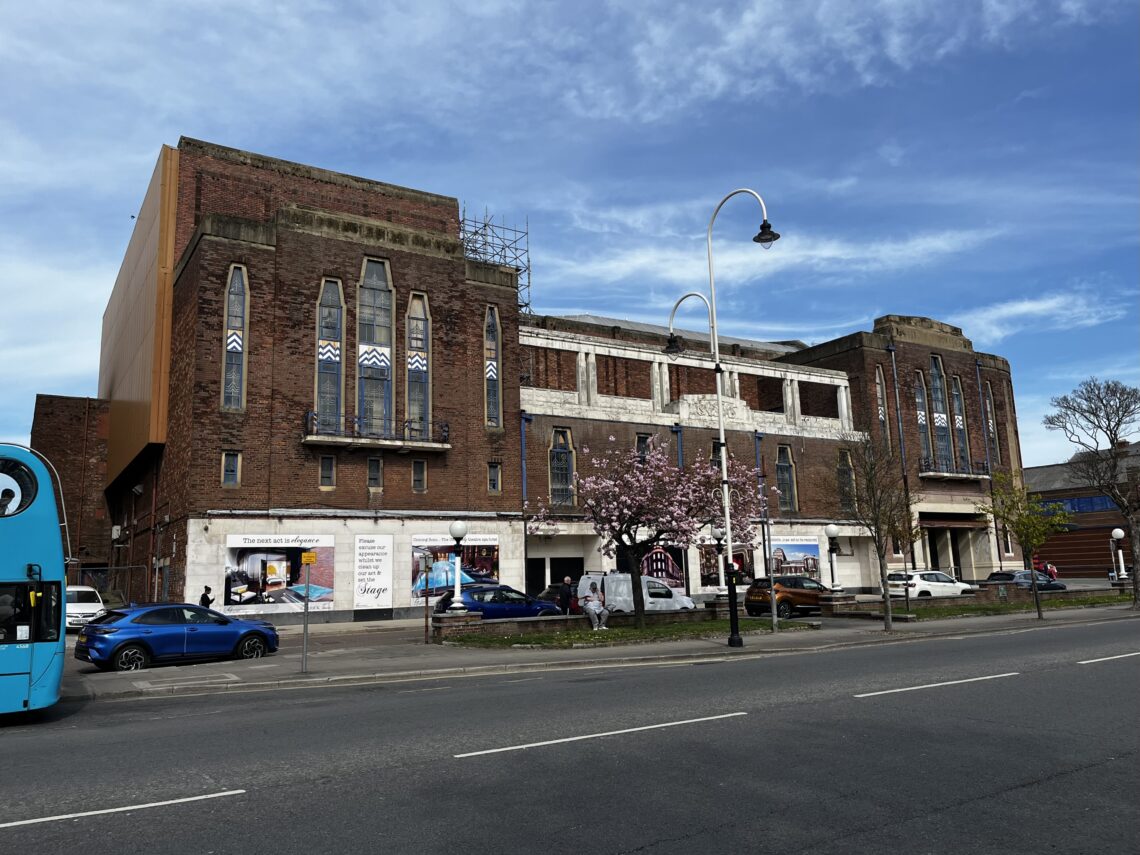 A large art deco building formed of two squareish shapes at the side with a lower main body in the middle. The building is in a bad state of repair, with hoarding and scaffolding around it.
