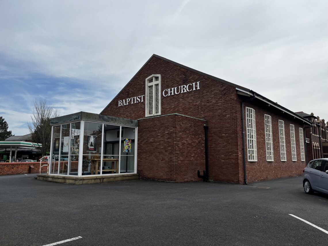Side/front view of a smallish church building, fairly plain, made out of brick. Side windows are made out of rectangular patterned concrete, there is large BAPTIST CHURCH lettering above the door, and there is also a small all-glass foyer at the front of the building.