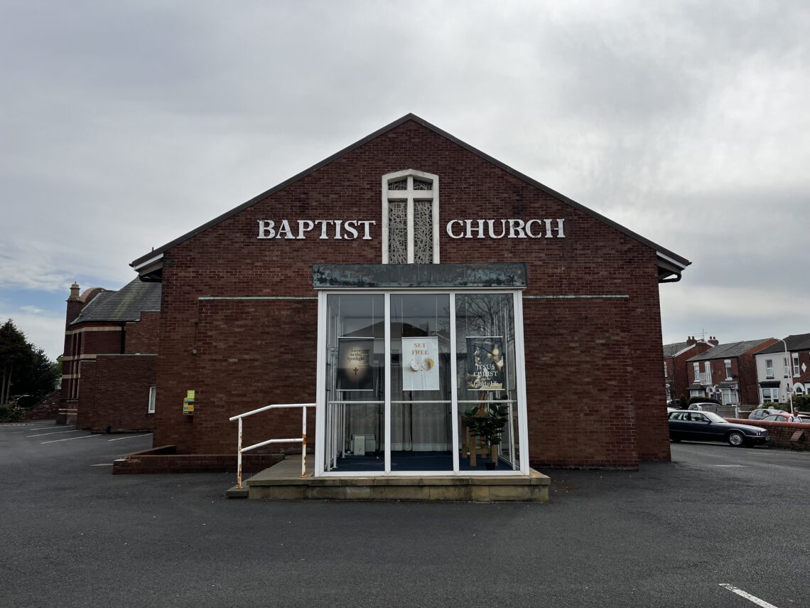 Front view of a plain brick church building, showing BAPTIST CHURCH lettering above the all-glass entrance foyer.